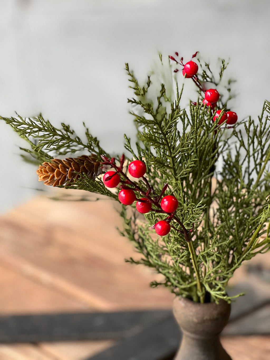 A festive artificial greenery arrangement featuring red berries, a pinecone, and evergreen branches in a rustic ceramic pot, set against a softly blurred background.
