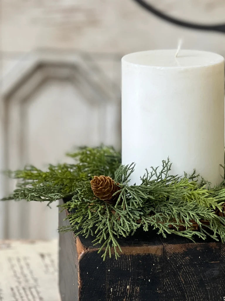 White candle on a wooden block with greenery and a pine cone.