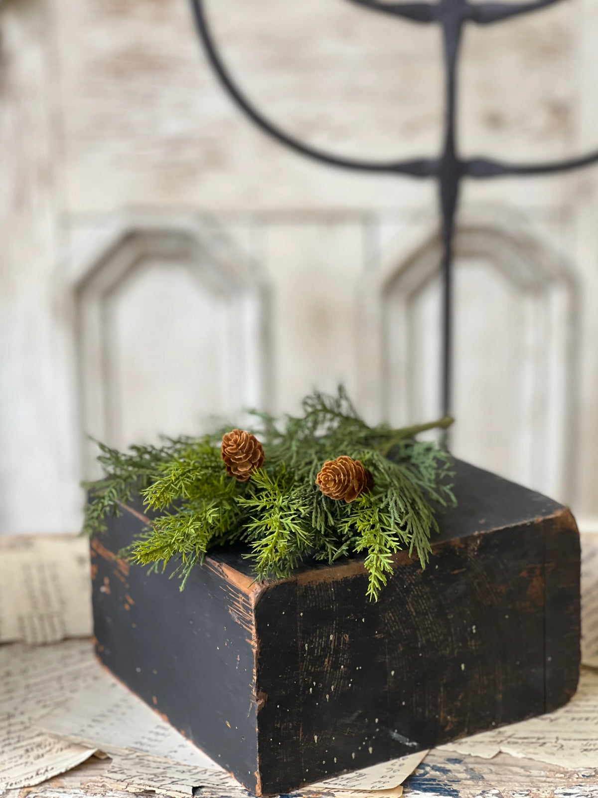 Close up of decorate greenery and pinecone stem  on a black block on a textured surface.