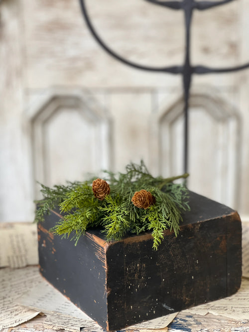 Close up of decorate greenery and pinecone stem  on a black block on a textured surface.