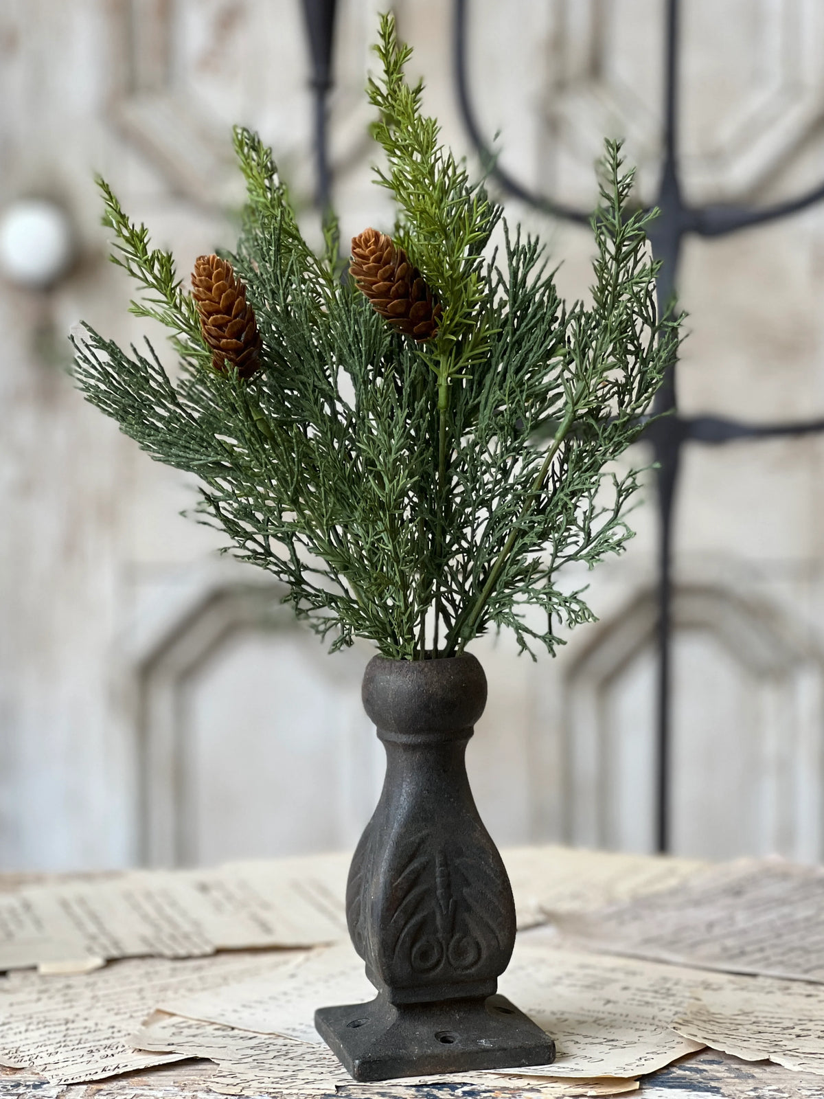 Decorative arrangement of greenery and pinecones in a black vase on a textured surface.