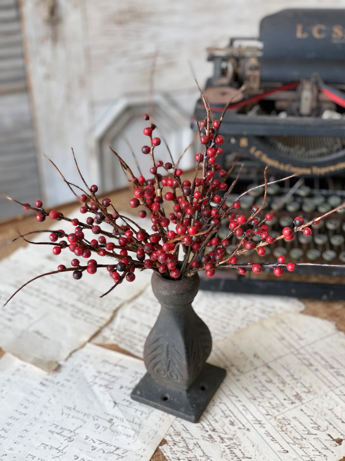 Decorative arrangement with red berried stem in a metal vase on a rustic surface with an old typewriter in the background.
