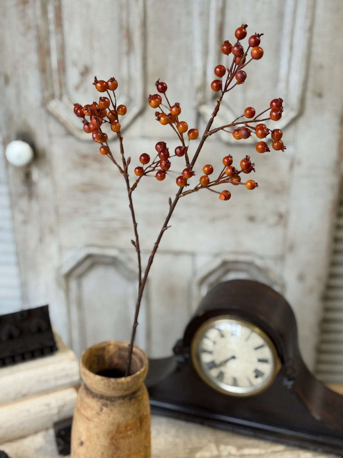 Decorative branch with red orange berries in a vase next to an old clock on a wooden surface.