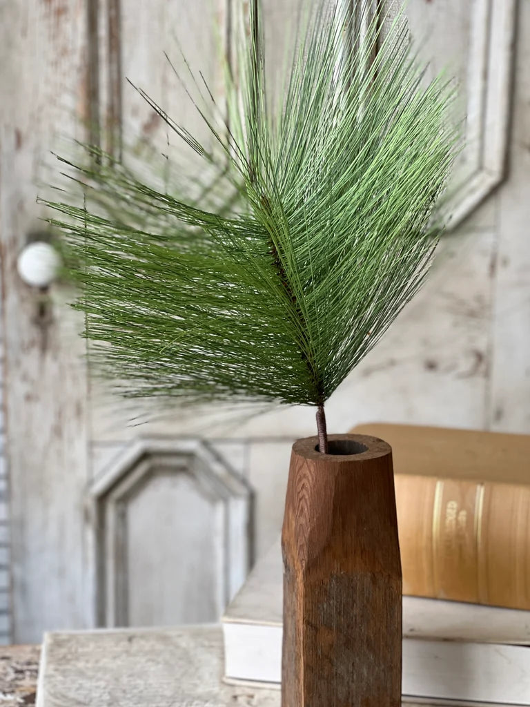 Green faux pine stem in a wooden vase against a rustic wall.