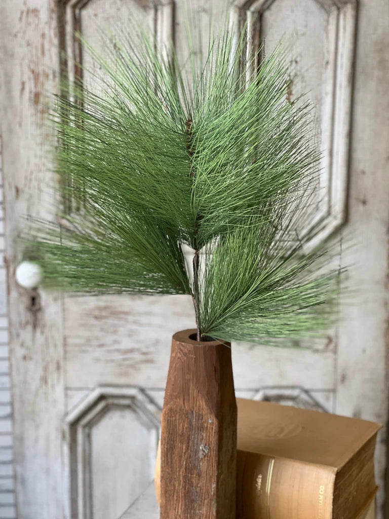 Close up of green faux pine stem in a wooden vase against a rustic wall.