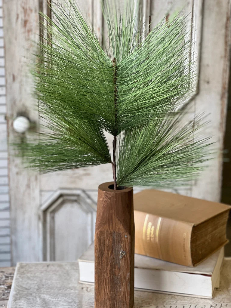 Green faux pine stem in a wooden vase against a rustic wall.