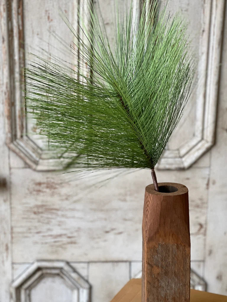 Close up of green faux pine stem in a wooden vase against a rustic wall.