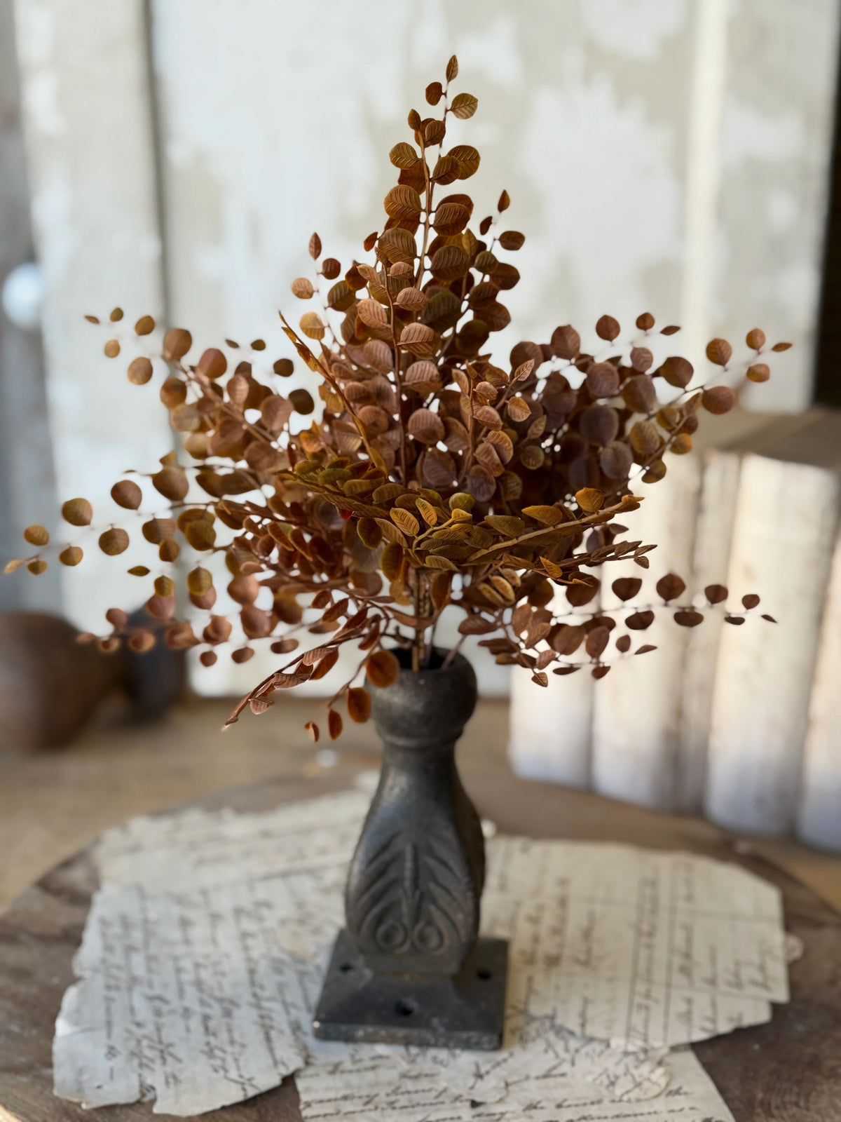 Decorative faux stem of dried copper brown leaves in a rustic metal vase on a wooden surface with a blurred background.