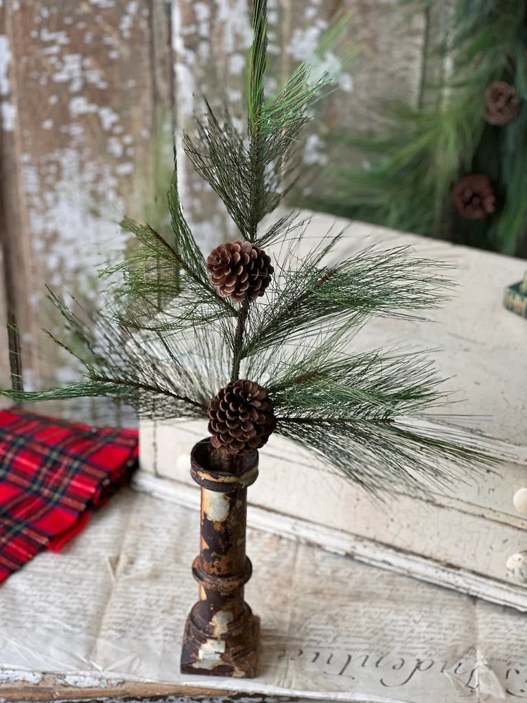 Decorative pine greenery stem with pinecones in a black pot on a rustic wooden surface.