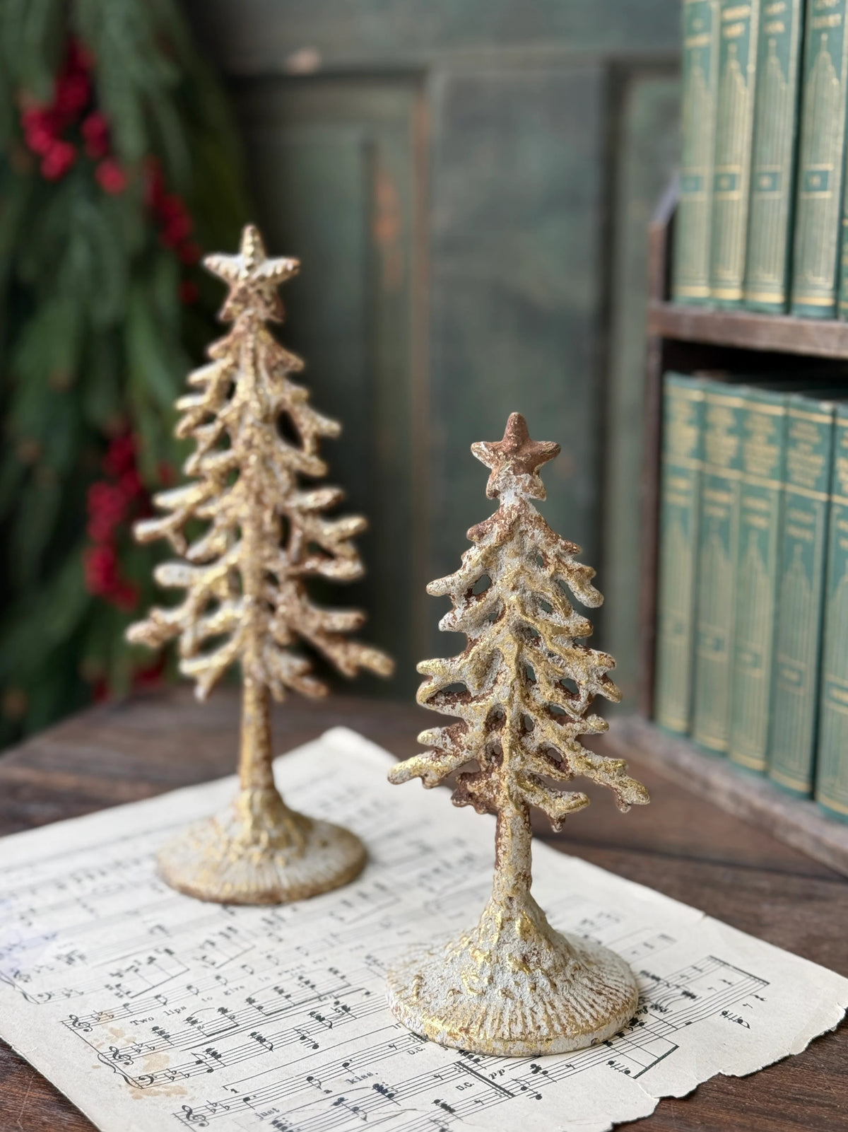 Two white washed gold metal decorative Christmas trees with stars on top, placed on sheet music, against a bookshelf background.