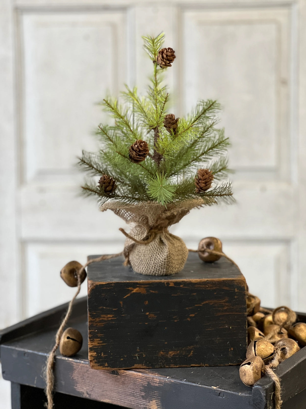 Small artificial Christmas tree with pinecones on a rustic wooden block against a white paneled wall.