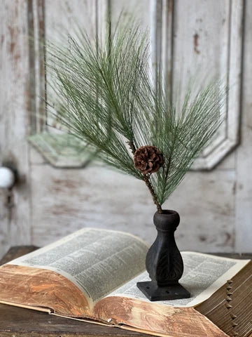 A pine stem with green pine needles and a small pinecone in a dark ceramic vase, styled on an open vintage book against a distressed white wooden door background.