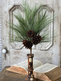 A rustic greenery stem featuring green pine needles and three brown pinecones arranged in a distressed metal candlestick holder, set on an open vintage book against a weathered white wooden door background.