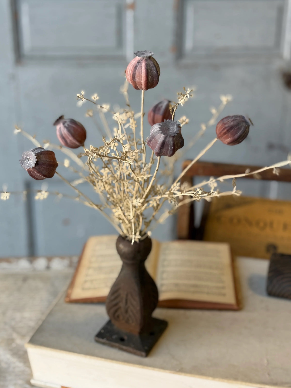 Decorative arrangement with dried flowers and pods on a metal stand, placed on an open book.