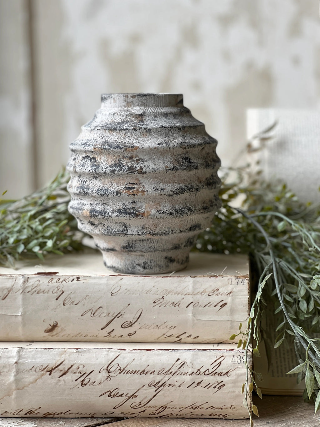 Textured ridged small wood vase with a distressed white wash finish over a dark bacground.  Vase is accented with greenery and sets on a stack of ivory colored books.