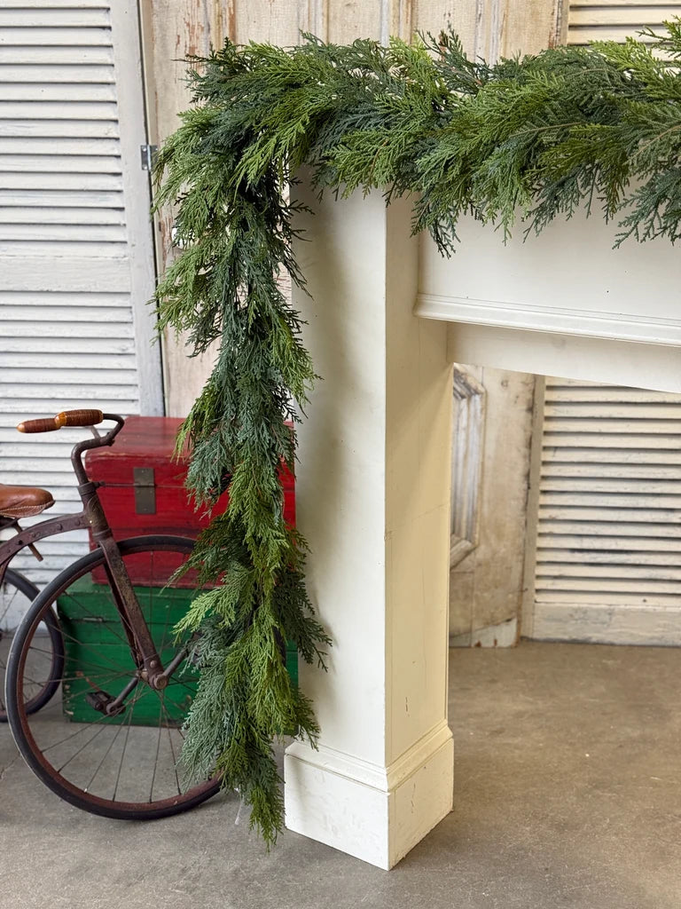 A green evergreen garland draped over a white fireplace mantel in a rustic indoor setting, with a vintage bicycle and red and green crates in the background.