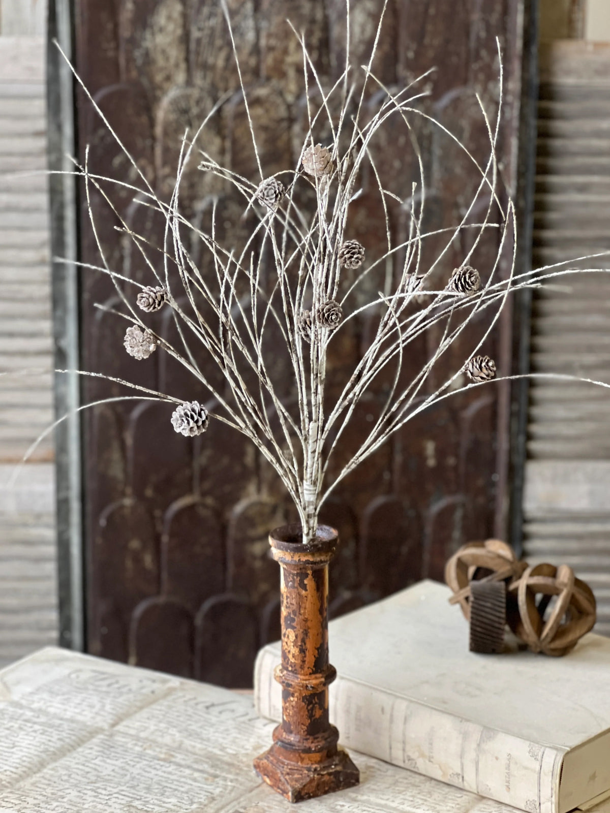 Decorative branch with pinecones on a rustic stand against a textured wall.