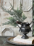 Decorative greenery cedar stem with a pine cone in an ornate black vase on a rustic surface.