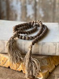 A strand of dark brown wooden beaded garland with two long beige jute tassels, draped over an old book on a rustic wooden surface.