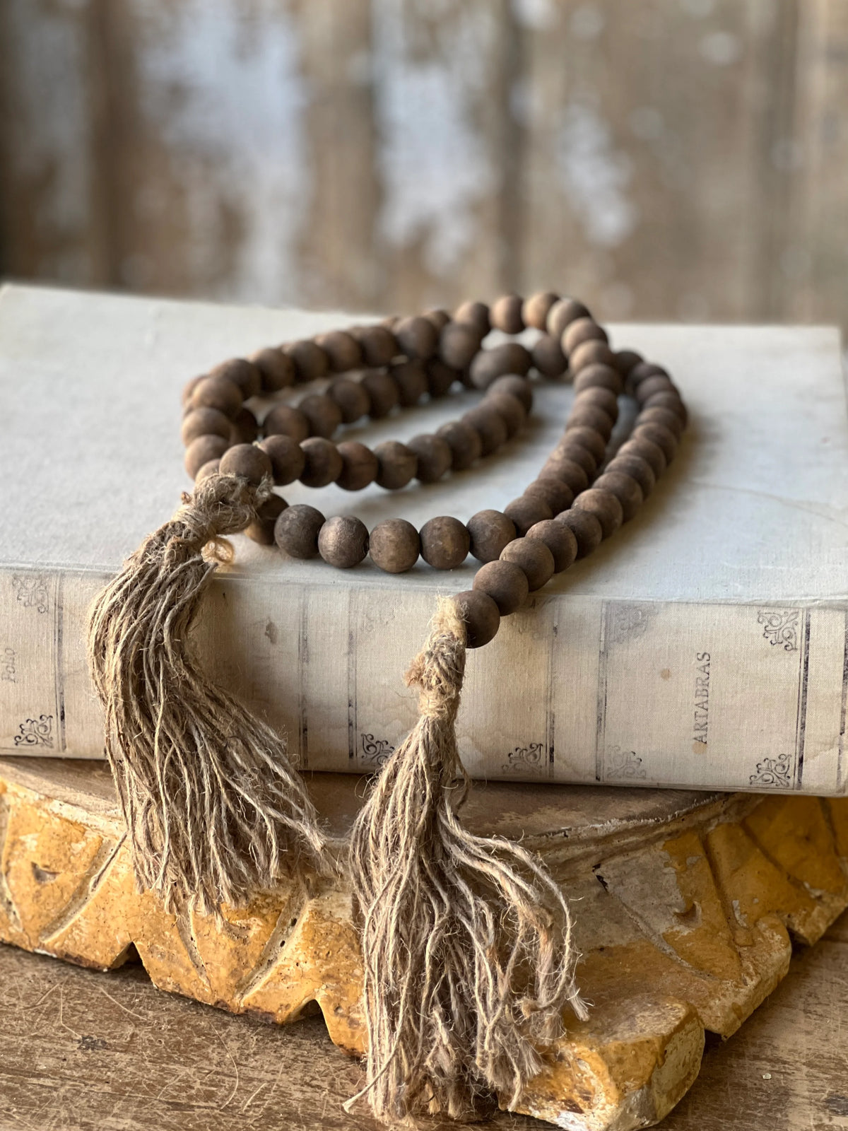 A strand of dark brown wooden beaded garland with two long beige jute tassels, draped over an old book on a rustic wooden surface.