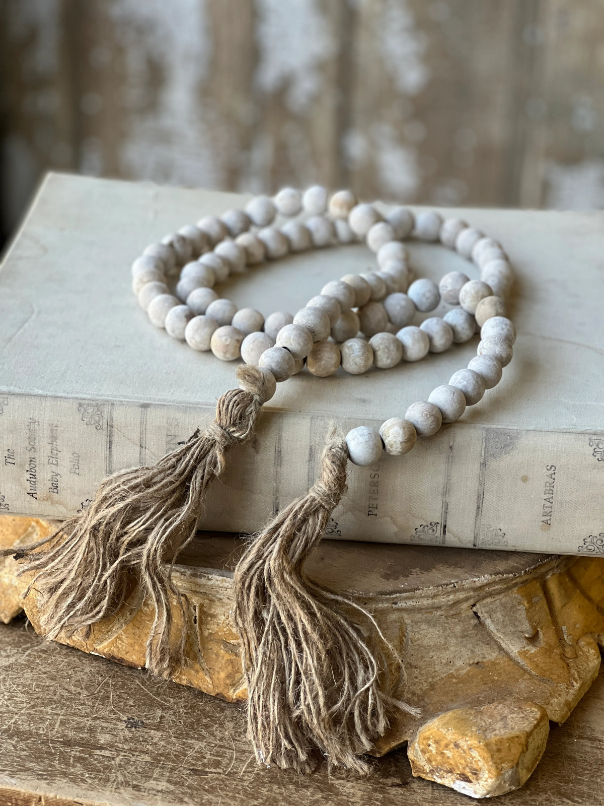 A beaded garland with round off-white beads and two long taupe tassel ends, draped over an old book on a rustic wooden surface.