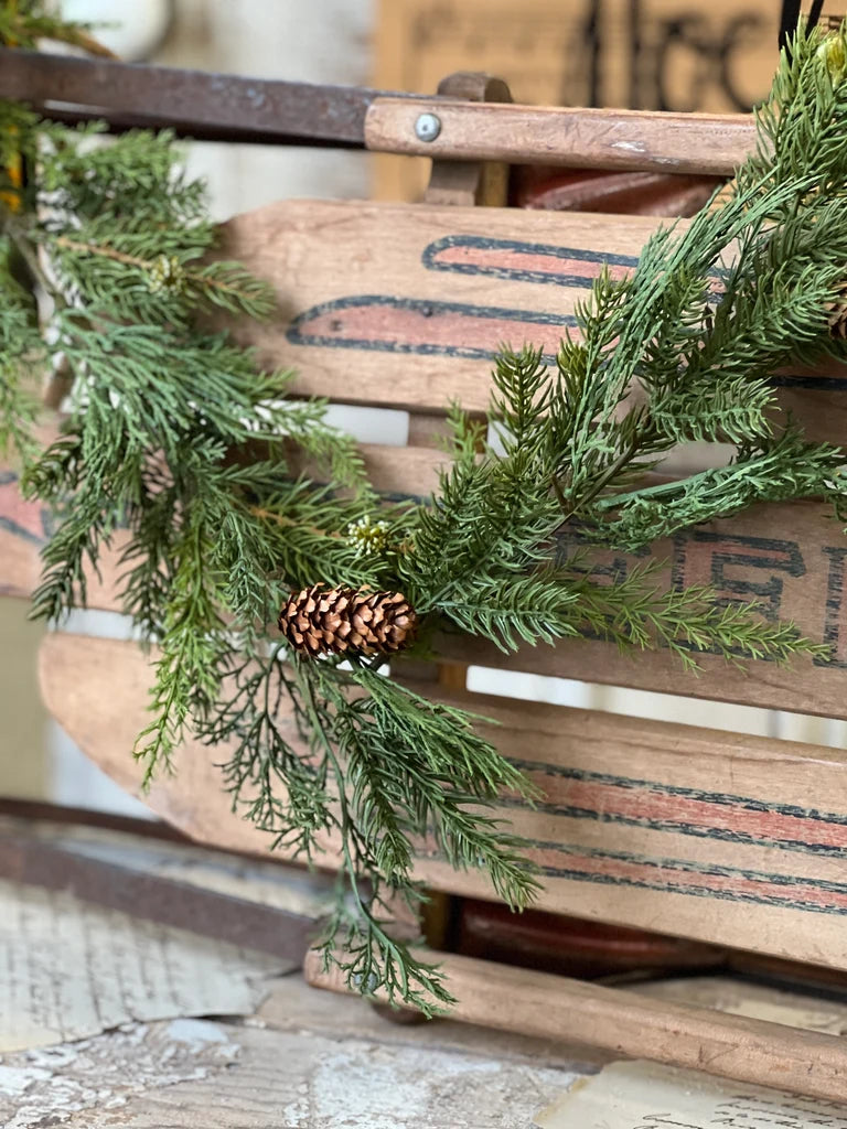 Green evergreen garland with pinecones draped over a rustic wooden sled or crate, photographed in a styled setting.