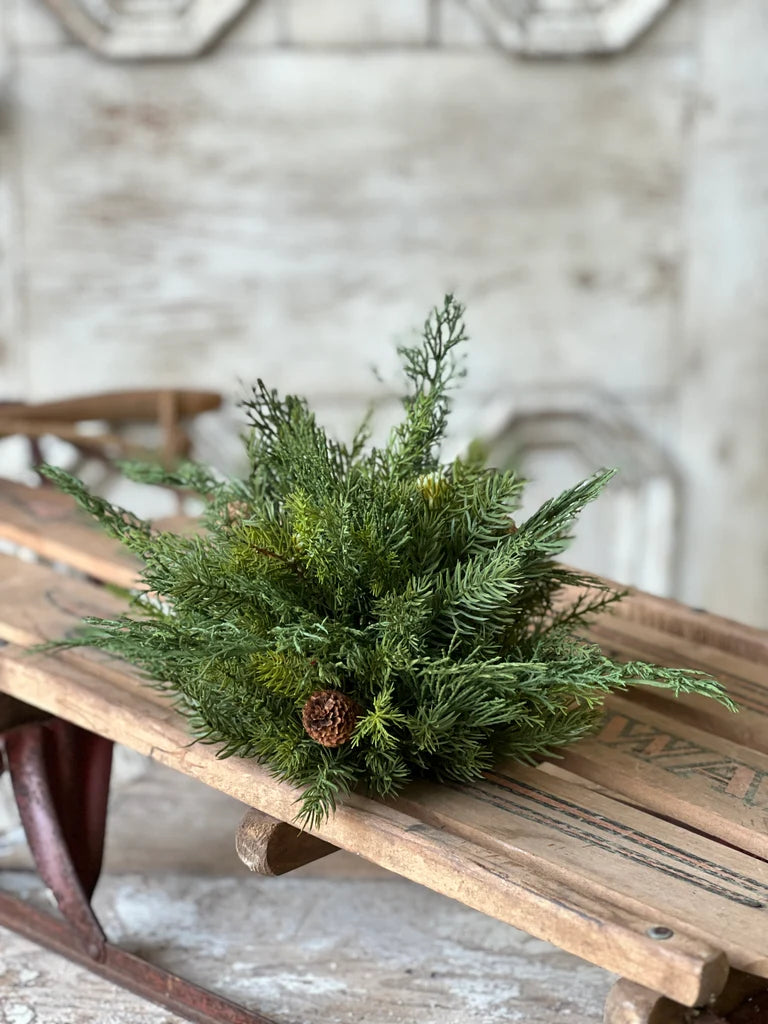 A green artificial pine half sphere with a small brown pinecone accent, displayed on a rustic wooden sled against a softly blurred, distressed white wall background.