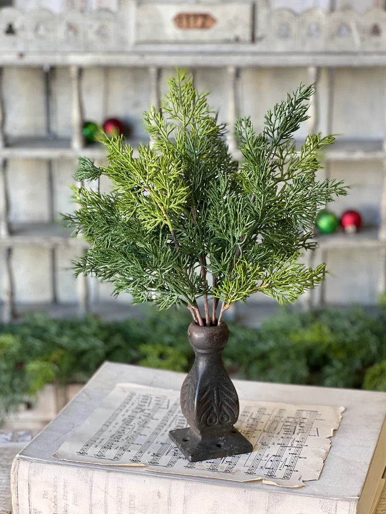 A green artificial pine or cypress-style branch arrangement in a dark brown carved pedestal vase, displayed on top of an old book with sheet music against a rustic shelving background.