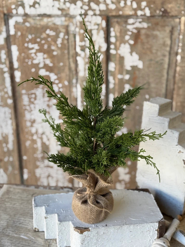 A small artificial cedar evergreen tree in a burlap-wrapped pot, displayed on a white decorative block against a rustic distressed wood background.