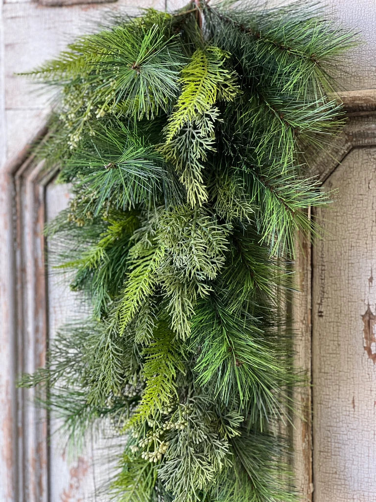 A green pine drop with mixed pine and fir branches hanging against a distressed white wooden door.