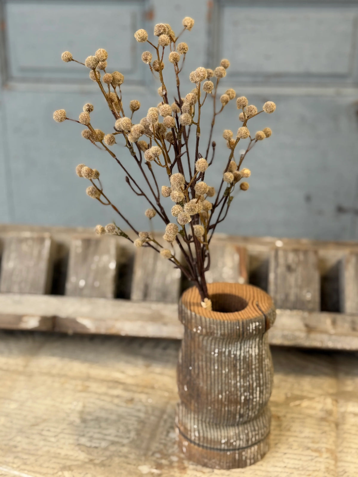 Dried floral stems in a rustic wooden vase on a wooden surface with a gray background.